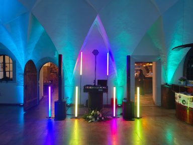 Bunte Lichtinstallation in einem Raum mit Bögen, DJ-Pult und Pflanzenarrangements. Photo by DJ Mirko Anders