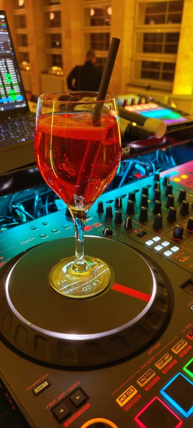 Ein Cocktail in einem Glas steht auf einem DJ-Pult mit bunten Lichtern im Hintergrund. Photo by DJ Mirko Anders