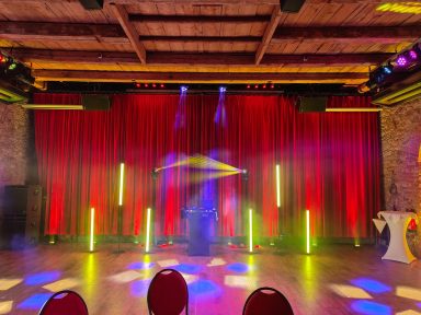 Bühne mit roten Vorhängen, bunten Lichtern und Nebel für eine Veranstaltung. Photo by DJ Mirko Anders