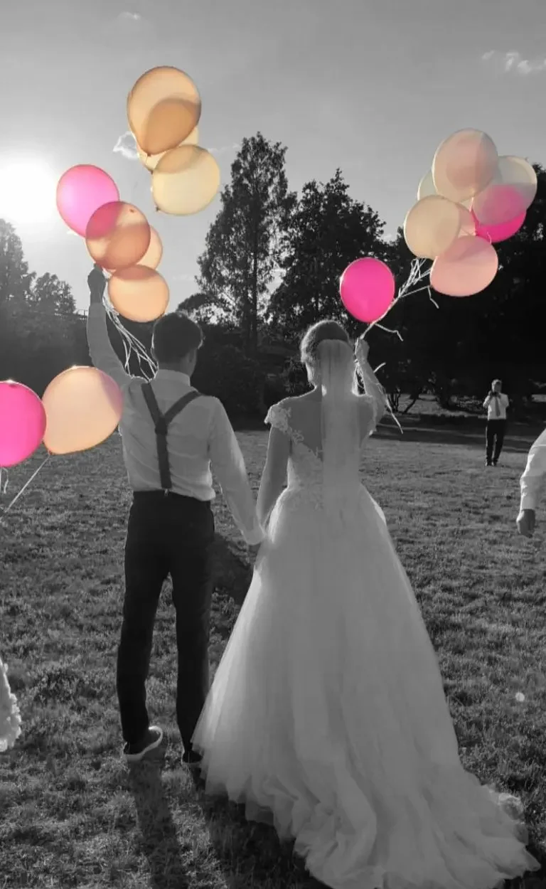 Hochzeitspaar hält bunten Ballons beim Sonnenuntergang auf einer Wiese.