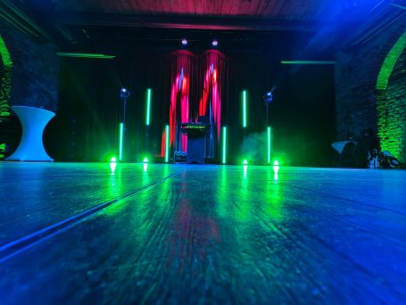 Bunte Lichter und beleuchtete Stangen in einem Eventraum mit Holzboden. Photo by DJ Mirko Anders