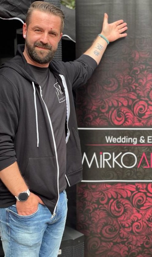 DJ Mirko Anders zeigt auf ein Werbeplakat für Hochzeitsevents.