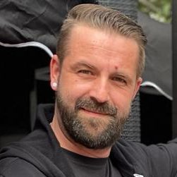 Ein Mann mit Bart lächelt, trägt ein schwarzes Oberteil und hat eine Ohrstecknadel.