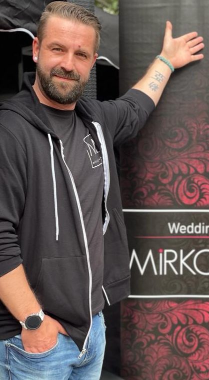 DJ Mirko Anders zeigt auf ein Werbeplakat für Hochzeitsevents.
