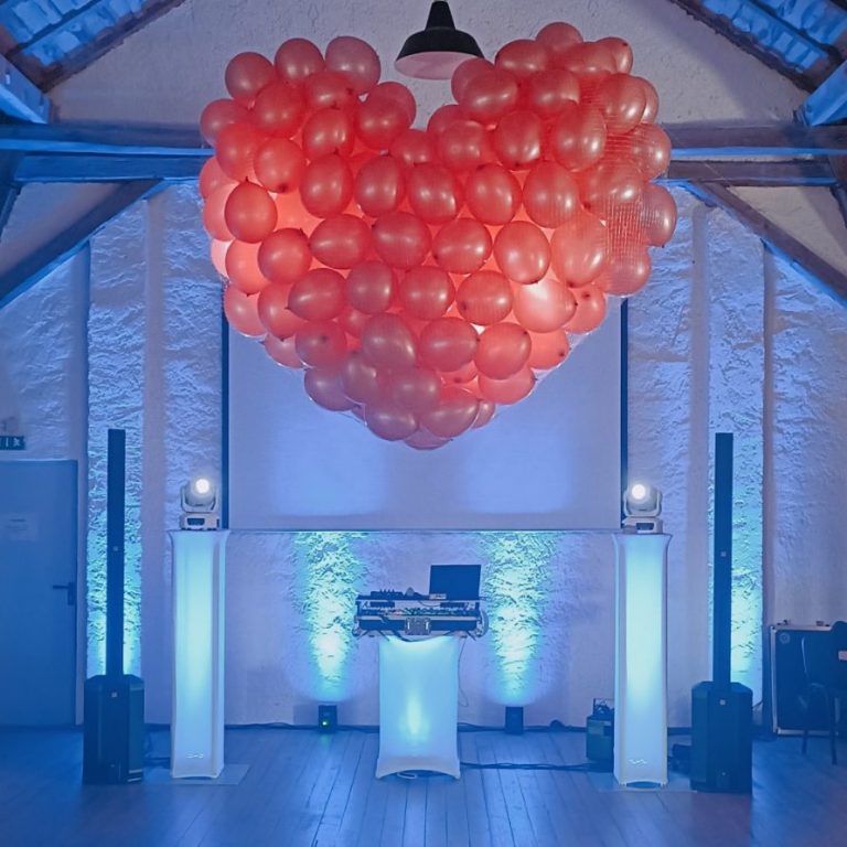 Rote Ballons in Herzform hängen über einer festlichen DJ-Bühne mit blauer Beleuchtung.